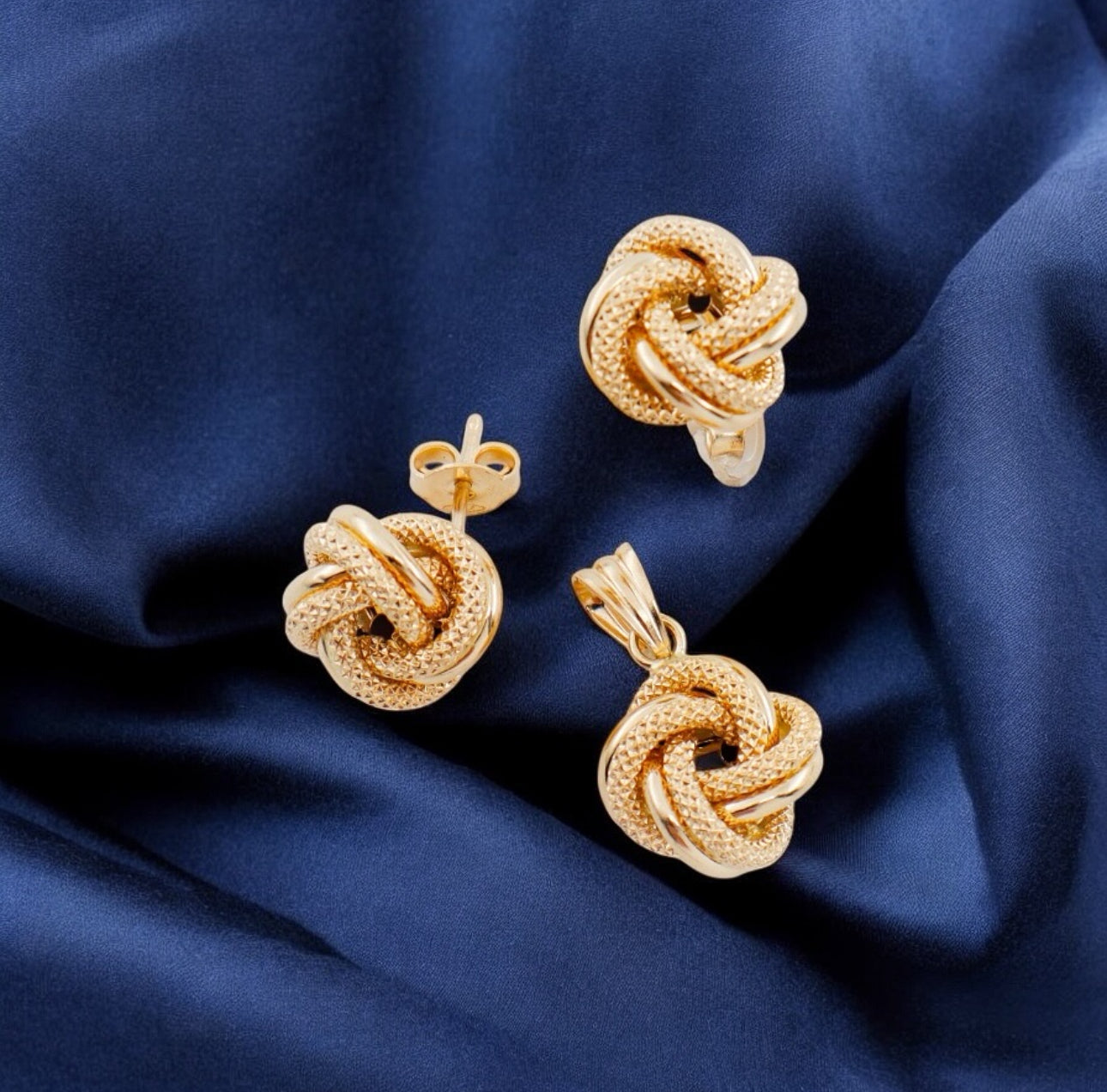 Nahaufnahme eines Schmucksets aus 18k Gelbgold auf dunkelblauem Satin. Der Anhänger in der Bildmitte sowie die beiden passenden Ohrstecker sind in Form kunstvoll verschlungener Goldknoten gestaltet. Die polierten und strukturierten Stränge reflektieren das Licht und erzeugen einen edlen Glanz.