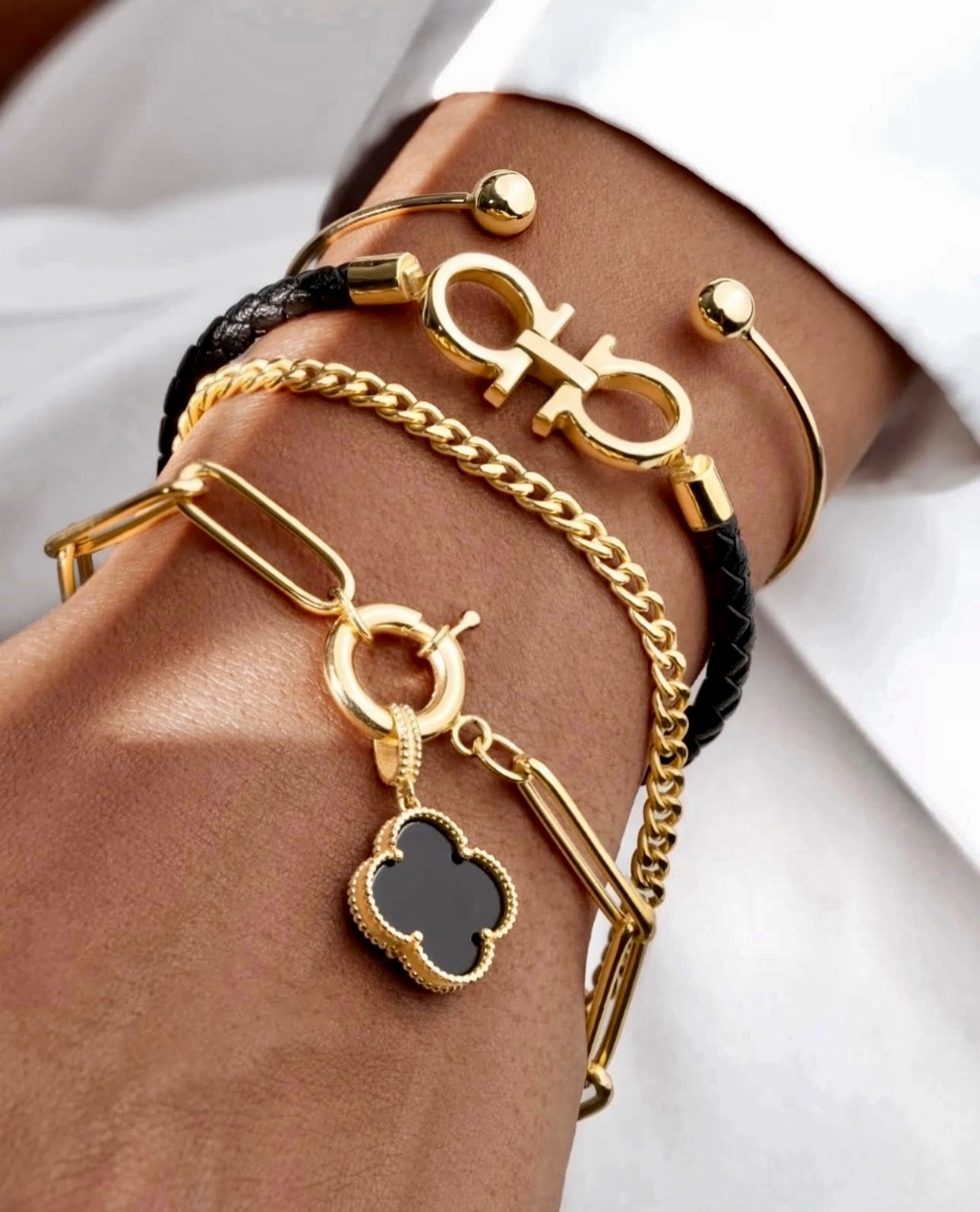 Nahaufnahme eines Handgelenks mit mehreren Armbändern, darunter der Anhänger „Barg-e Onyx“ aus 18 Karat Gelbgold – Kleeblattform mit schwarzer Onyx-Einlage, eingefasst in eine feine goldene Umrandung.