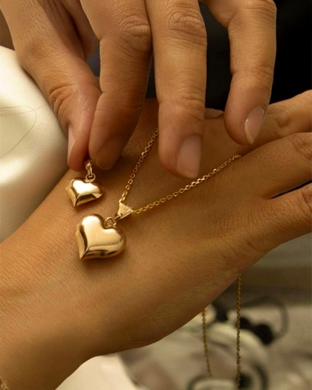 Zwei glänzende, herzförmige Anhänger aus 18k Gelbgold auf feiner Kette – in einer Hand gehalten, vor warmem, sanftem Hintergrund.