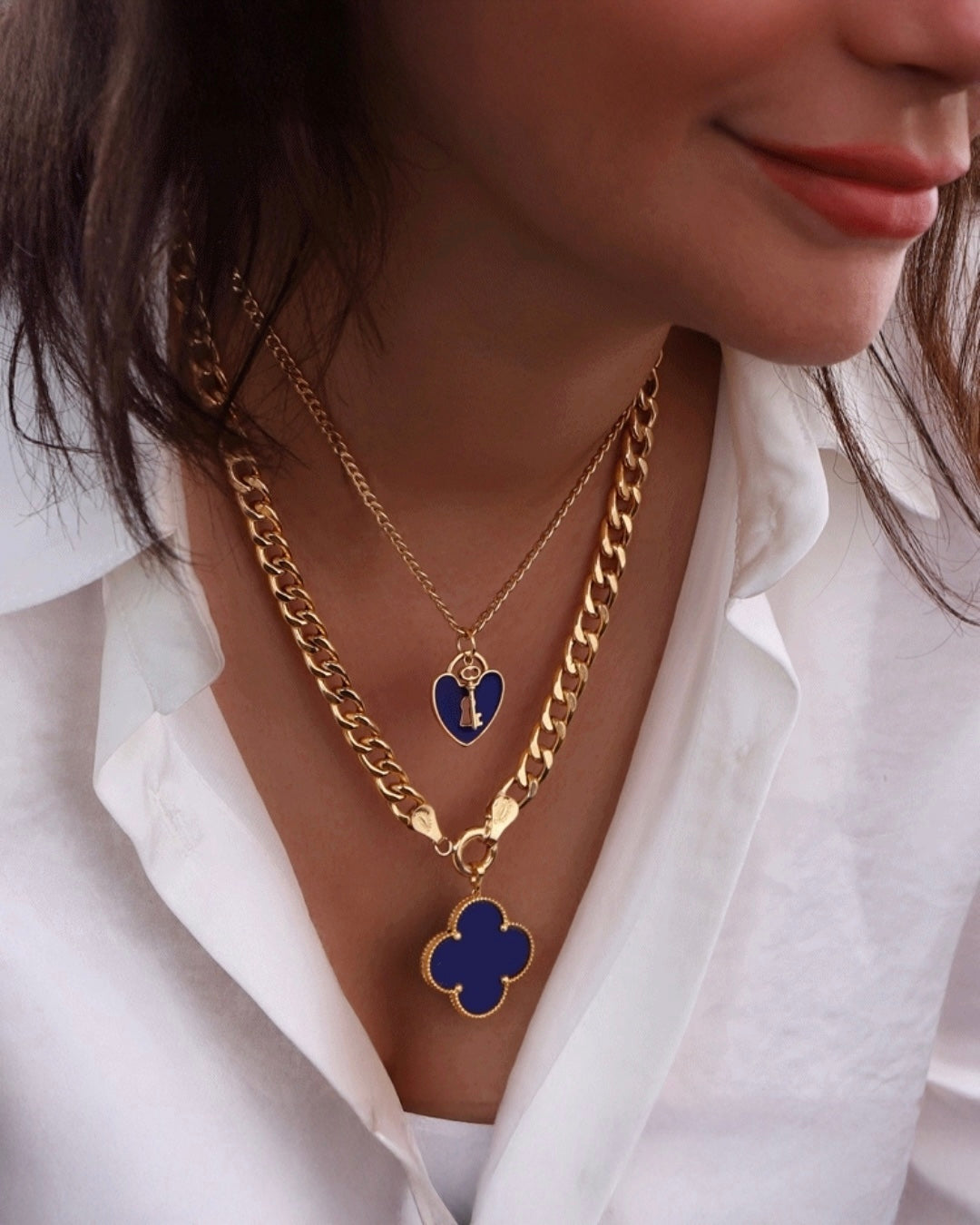 Frau trägt zwei goldene Halsketten aus 18 Karat – eine mit einem blauen Herzanhänger und Schlüsselmotiv, die andere mit einem großen, blauen Kleeblattanhänger. Die Ketten liegen übereinander auf einer weißen Bluse und verleihen dem Look eine elegante, moderne Note.