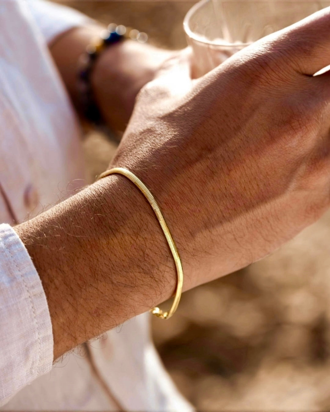 Feines, minimalistisches Armband aus 18k Gelbgold am Handgelenk eines Mannes, warmes Sonnenlicht, elegante Alltagsaufnahme mit natürlicher Hautstruktur und weichem Hintergrund.