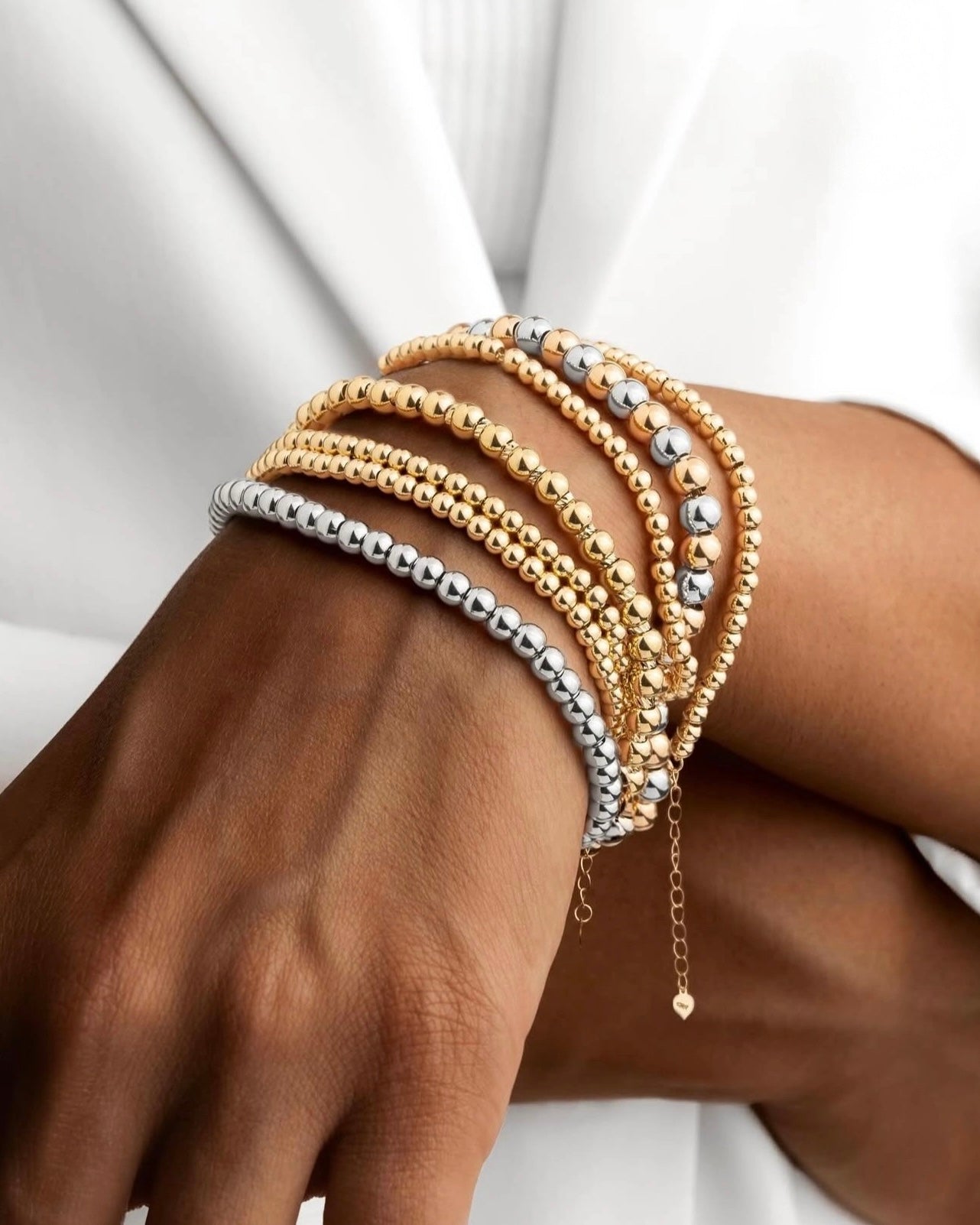 Mehrere Armbänder aus 18 Karat Gold und Weißgold in Kugeldesign, elegant am Handgelenk getragen und kunstvoll übereinander gestapelt – moderner, luxuriöser Look in Kombination mit weißem Outfit.