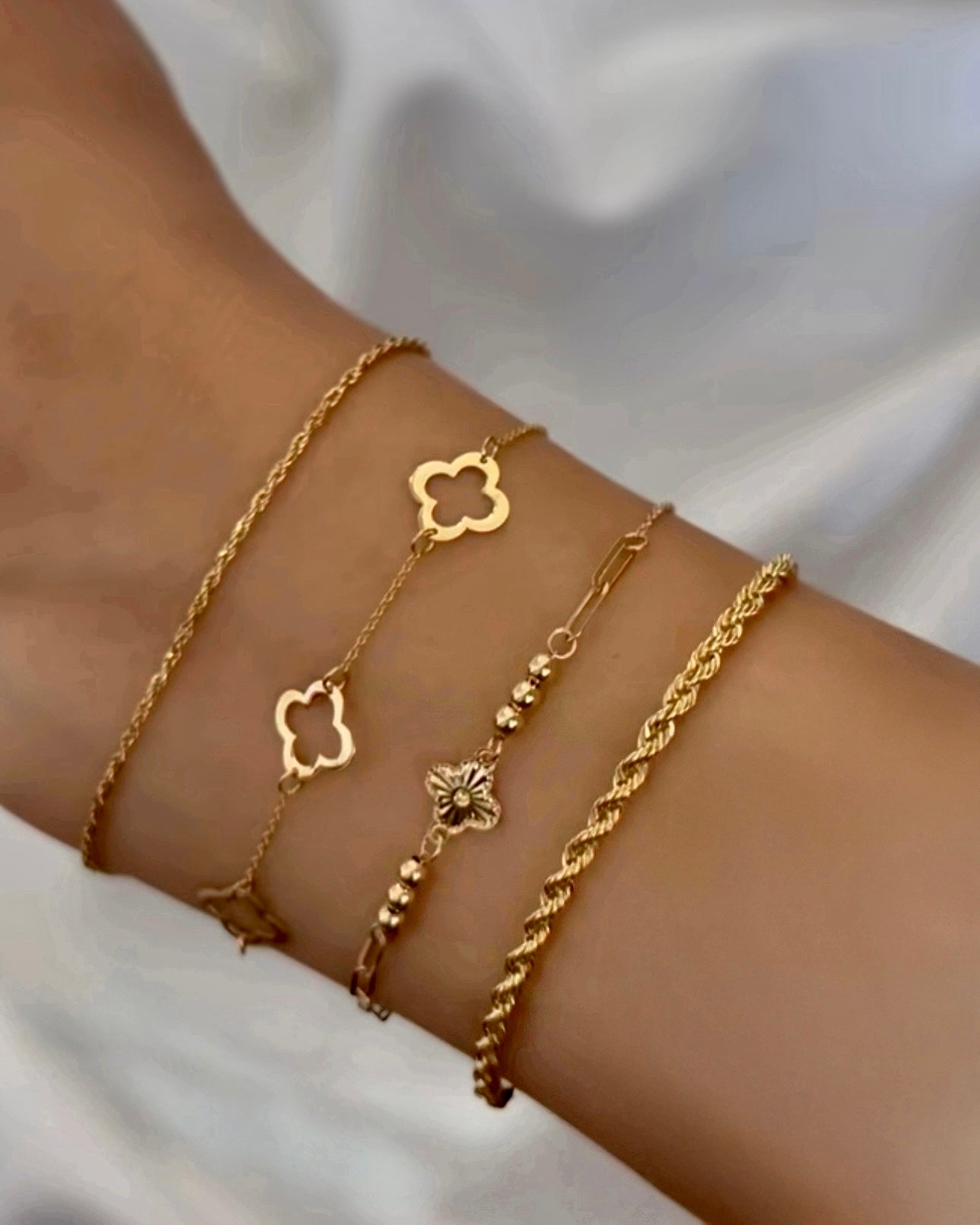 Vier filigrane Armbänder aus 18 Karat Gelbgold, kombiniert am Handgelenk – Designs mit Kleeblatt-, Schmetterlings- und Kordelelementen, teils mit kleinen goldenen Kügelchen, elegant und harmonisch zusammengestellt.