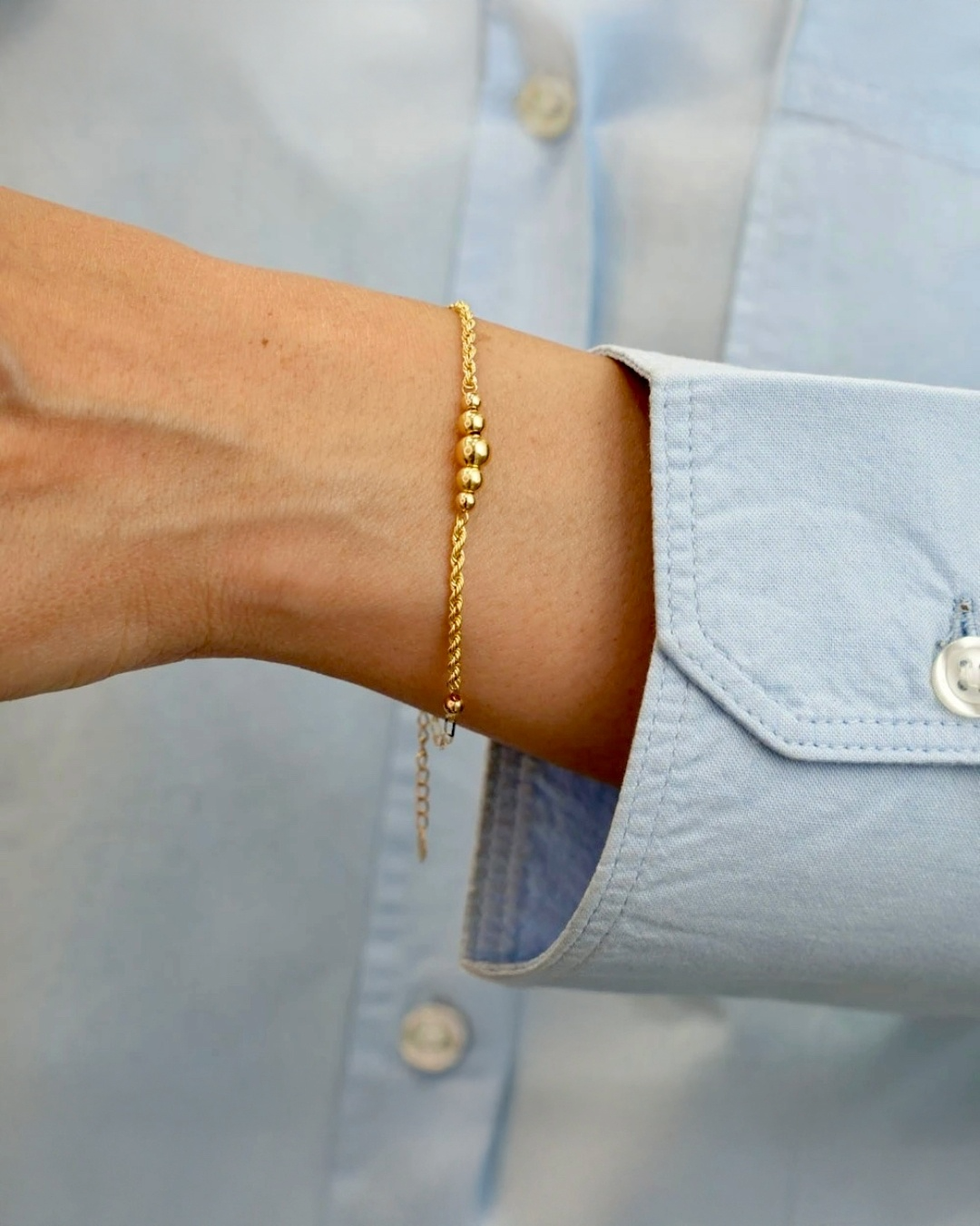 Feines 18k Goldarmband mit kleinen polierten Goldkugeln, getragen am Handgelenk einer Person mit hellblauem Hemd; zartes, elegantes Design mit filigraner Kette.