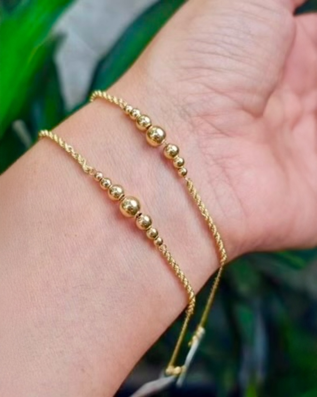 Zwei filigrane 18k-Goldarmbänder mit kleinen, polierten Goldkugeln liegen übereinander am Handgelenk. Die Armbänder haben eine zarte Kordelstruktur, die Kugeln variieren in der Größe. Hintergrund leicht unscharf mit grünen Pflanzen.