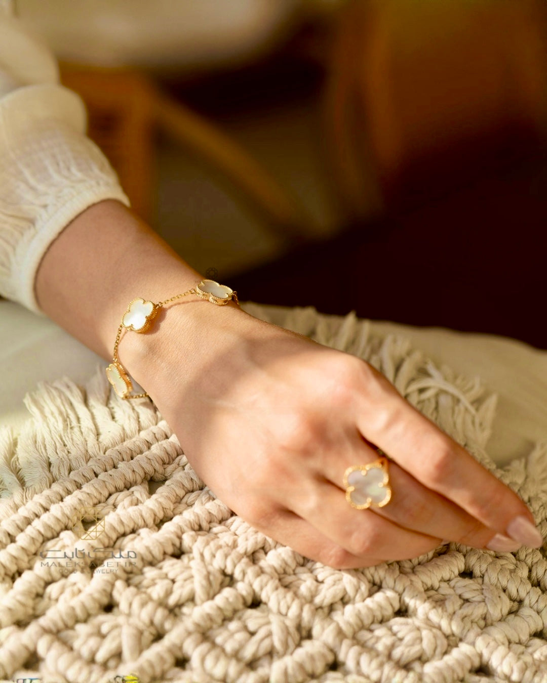 Hand mit elegantem 18k-Gelbgold-Armband und passendem Ring mit weißem Stein in Kleeblatt-Form, warmes Licht, luxuriöse Schmuckfotografie.