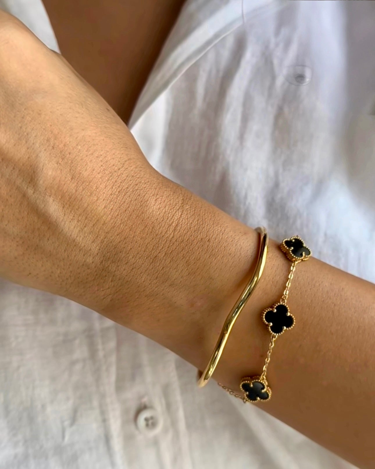 Ansicht eines Handgelenks mit zwei Armbändern: einem schlichten, geschwungenen Armreif aus Gelbgold und einem filigranen Goldarmband mit schwarzen, vierblättrigen Onyx-Elementen, getragen zu einer weißen Bluse.
