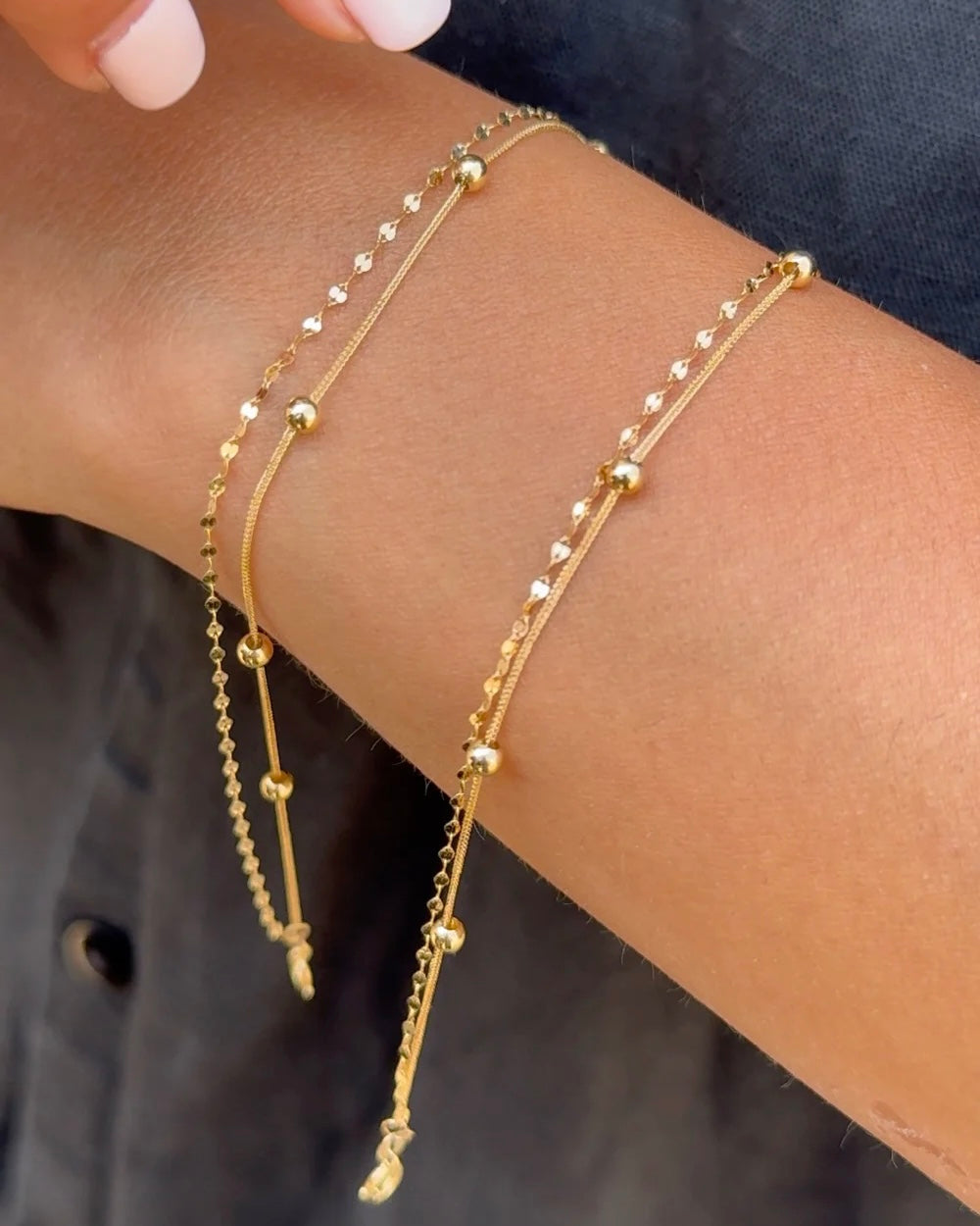 Zwei filigrane Armbänder aus 18 Karat Gelbgold, verziert mit kleinen, glänzenden Kügelchen und feinen Gliedern – elegant am Handgelenk getragen, kombiniert mit einem dunklen Outfit für einen schlichten, edlen Look.