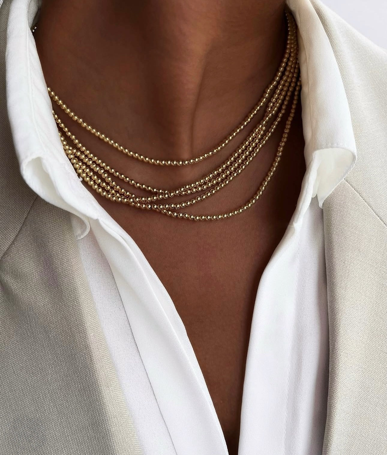 Elegante, mehrreihige Kette aus hochglanzpolierten Goldkügelchen, die eng am Hals anliegt. Das Design verbindet Schlichtheit mit luxuriöser Raffinesse und wirkt besonders edel in Kombination mit einem weißen Hemd und hellem Blazer.