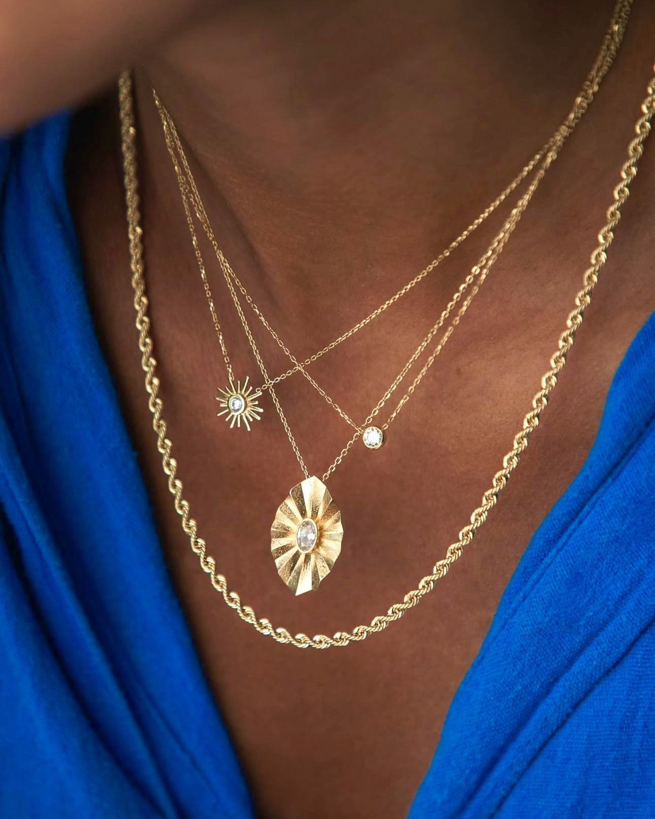 Mehrlagige Halsketten aus 18 Karat Gelbgold – kombiniert aus einer gedrehten Kordelkette und feinen Ketten mit Anhängern, darunter eine Sonne mit Zirkonia, ein runder Stein und ein kunstvoll gearbeiteter Anhänger, getragen zu einem blauen Oberteil.
