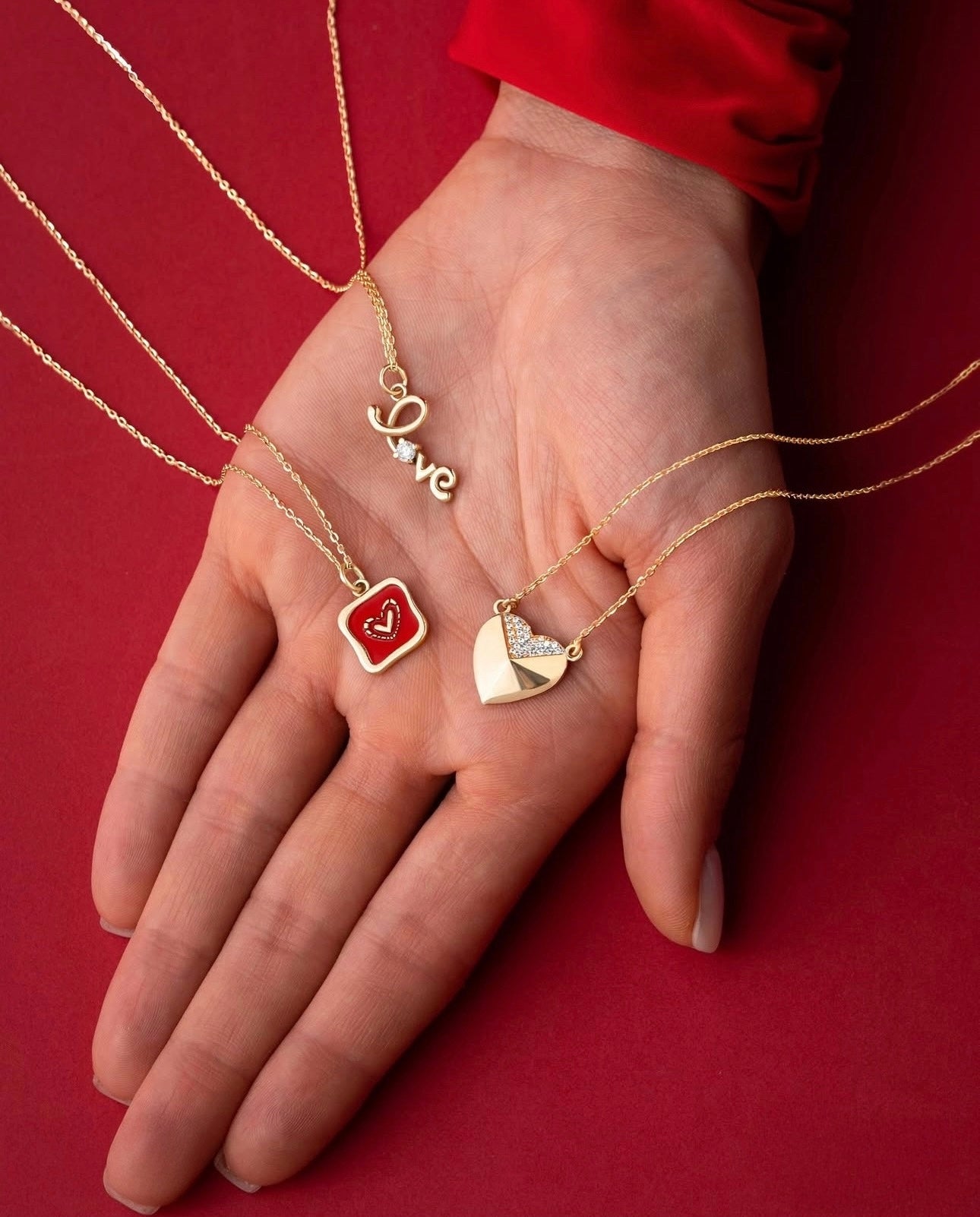 Nahaufnahme einer Hand mit drei filigranen 18k Gelbgold-Halsketten auf rotem Hintergrund, jede mit einem romantischen Anhänger: Schriftzug „Love“ mit kleinem Stein, rotes Emaille-Herz im Quadrat und goldenes Herz mit teils pavégefassten Steinen.