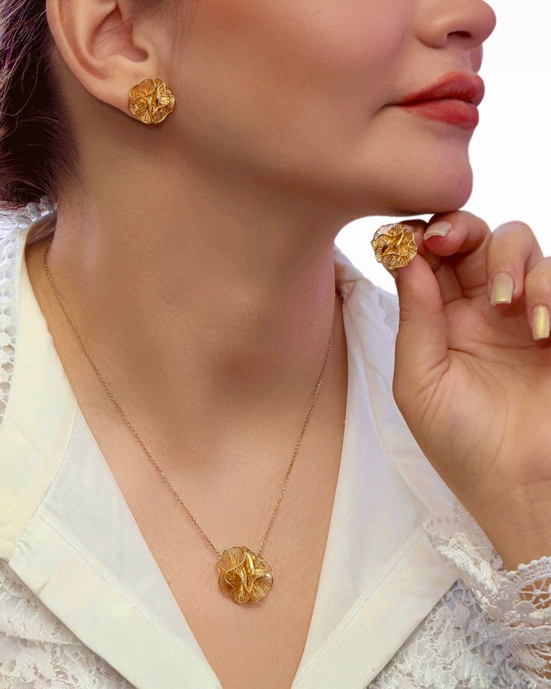 Frau mit goldfarbenen, organisch geformten Ohrringen und passender Halskette. Sie hält einen der Ohrringe zwischen den Fingern, wodurch die strukturierte, blütenartige Oberfläche gut sichtbar ist. Sie trägt eine weisse, leicht strukturierte Bluse; Hintergrund neutral und hell, Fokus klar auf dem Schmuck.