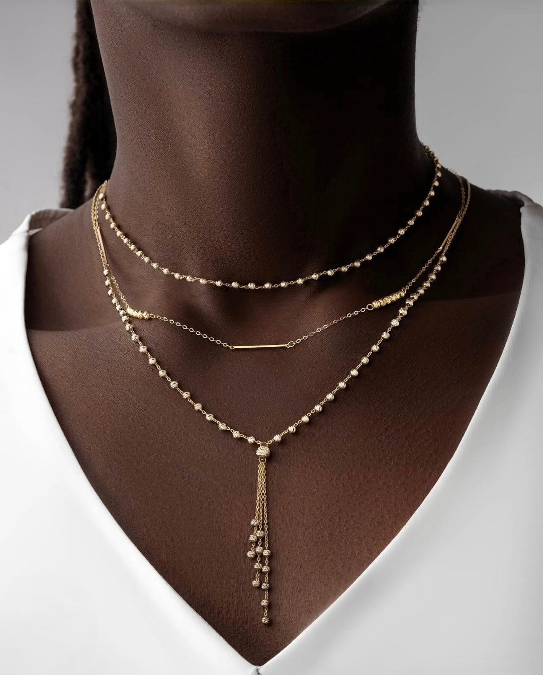 Nahaufnahme eines eleganten, dreilagigen 18k Gelbgold-Halsketten-Ensembles auf dunkler Haut. Die obere Kette besteht aus kleinen, polierten Goldkügelchen, die mittlere aus einer feinen Gliederkette mit schlichtem Goldstab, und die untere Kette aus filigranen Goldperlen mit einem langen, herabfallenden Anhänger aus zarten Goldketten. Das Ensemble wirkt luxuriös, modern und perfekt abgestimmt auf das weiße Oberteil.