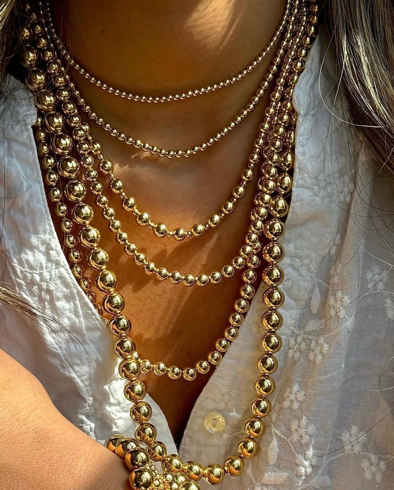 Nahaufnahme mehrerer übereinander getragener Halsketten aus 18k Gelbgold, bestehend aus unterschiedlich großen, hochglanzpolierten Goldkugeln („Morvarid-e Shahanshahi“ – Goldkugeln der Könige). Die Ketten variieren in Länge und Kugeldurchmesser, was ein luxuriöses, mehrlagiges Statement-Look erzeugt. Getragen zu einer weißen Bluse mit feiner Stickerei.