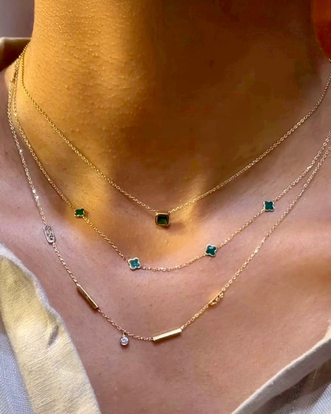 Nahaufnahme eines Halses mit drei filigranen Halsketten aus 18k Gelbgold. Die mittlere Kette („Peymân-e Sabz“ – Das grüne Versprechen) ist mit mehreren kleinen, kleeblattförmigen Anhängern in sattem Smaragdgrün besetzt. Darüber eine kurze Kette mit einem einzelnen quadratischen grünen Edelsteinanhänger, darunter eine längere Kette mit schlichten Goldzylindern und einem einzelnen klaren Steinanhänger. Getragen zu einem hellen, tief ausgeschnittenen Oberteil.