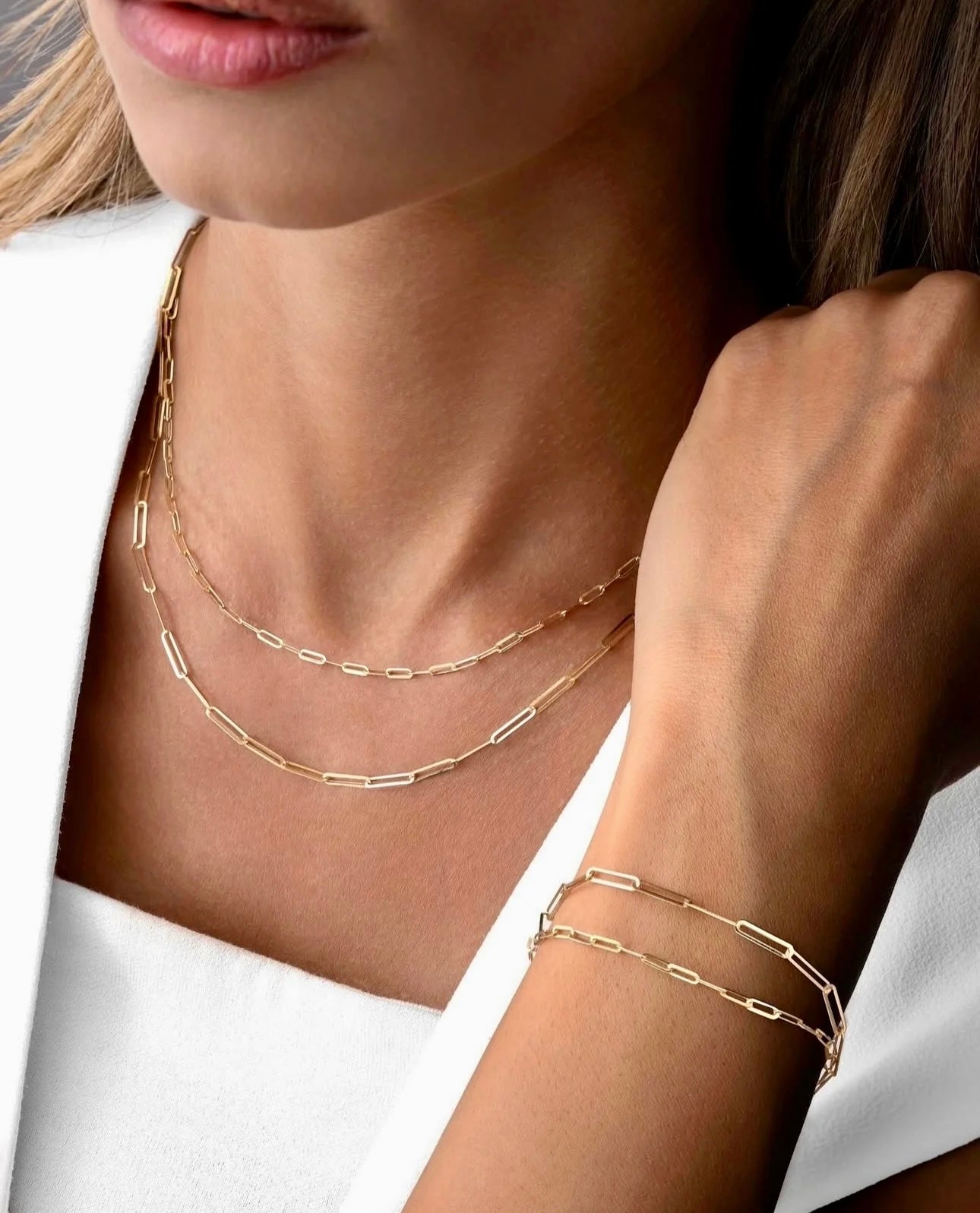 Nahaufnahme einer Frau in weißer Kleidung, die zwei zarte 18k-Gelbgold-Halsketten und ein passendes Armband mit rechteckigen Gliedern trägt. Der Fokus liegt auf dem eleganten, minimalistischen Goldschmuck im Layering-Stil.