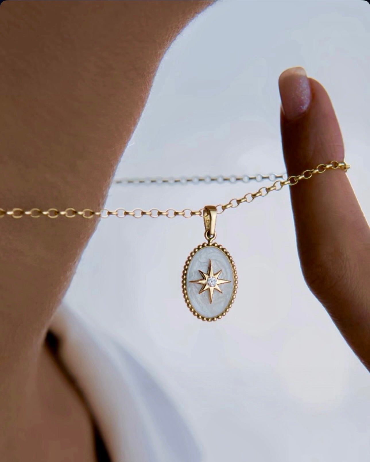 Nahaufnahme einer Halskette aus 18 Karat Gelbgold mit ovalem Anhänger – Perlmuttgrund und ein goldener Stern mit funkelndem Stein in der Mitte, elegant zwischen den Fingern gehalten und vor hellem Hintergrund präsentiert.