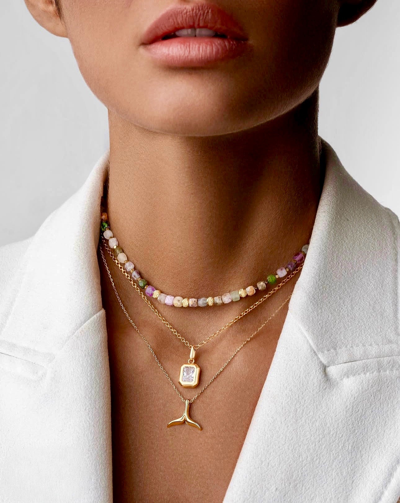 Drei übereinander getragene Halsketten: kurze bunte Edelstein-Perlenkette, feine Goldkette mit rechteckigem facettiertem Anhänger, darunter goldener Flossen-Anhänger; getragen zu weißem Blazer.