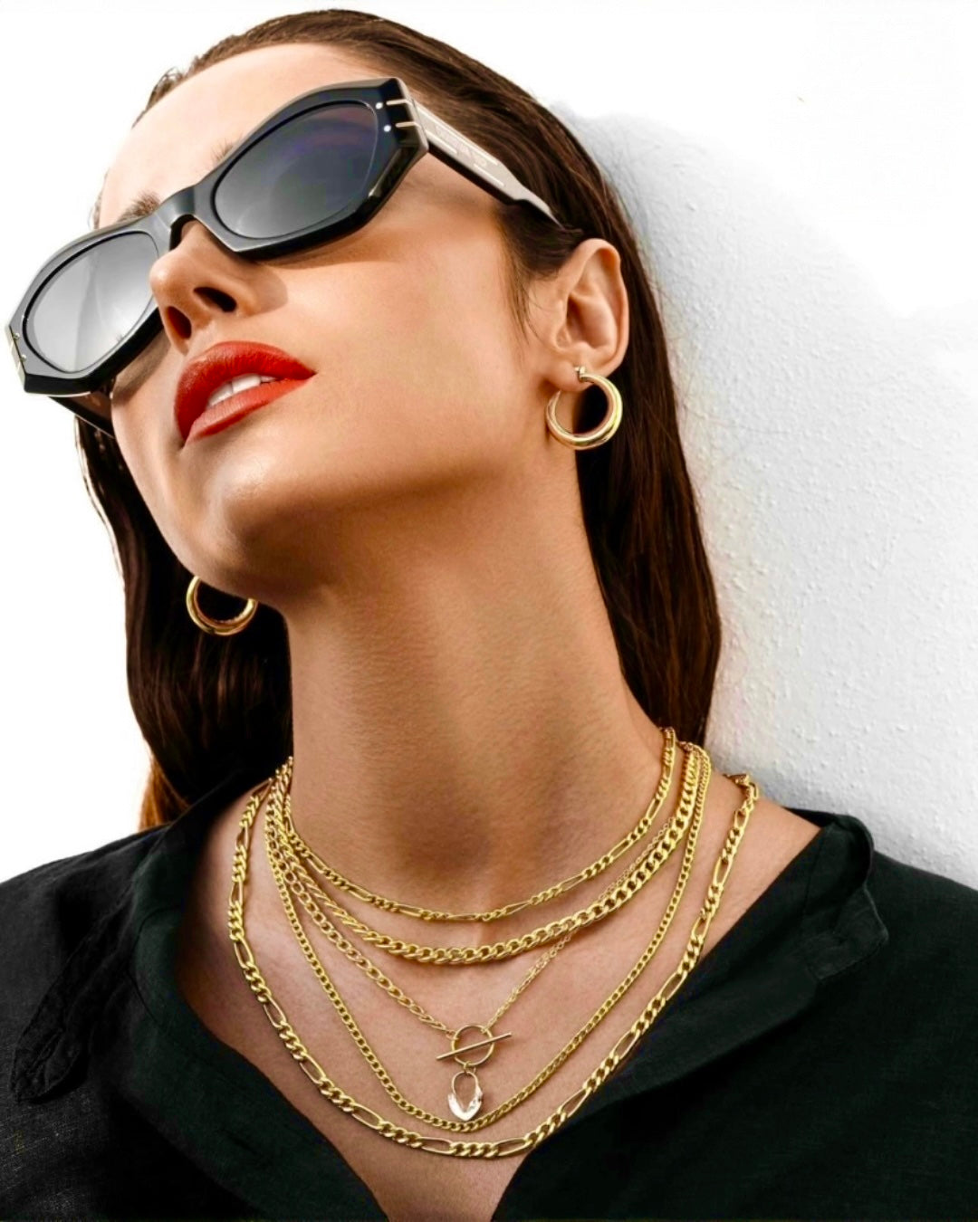 Elegantes Model mit schwarzer Sonnenbrille, rotem Lippenstift und mehreren übereinander getragenen Goldketten sowie goldenen Creolen, stilvoll vor hellem Hintergrund inszeniert.