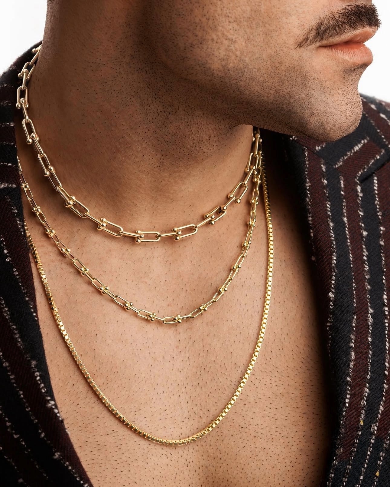 Nahaufnahme einer 18k Gelbgold-Halskette im Venice-Design, getragen von einem Mann in offenem, gestreiftem Sakko, kombiniert mit zwei weiteren Goldketten in unterschiedlichen Stilen.