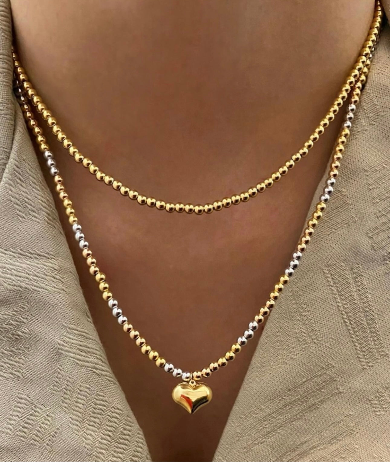 Doppelreihige Halskette aus 18k Gelb- und Weißgold mit polierten Goldperlen, untere Kette verziert mit einem glänzenden Herzanhänger, getragen zu einem beigen V-Ausschnitt.
