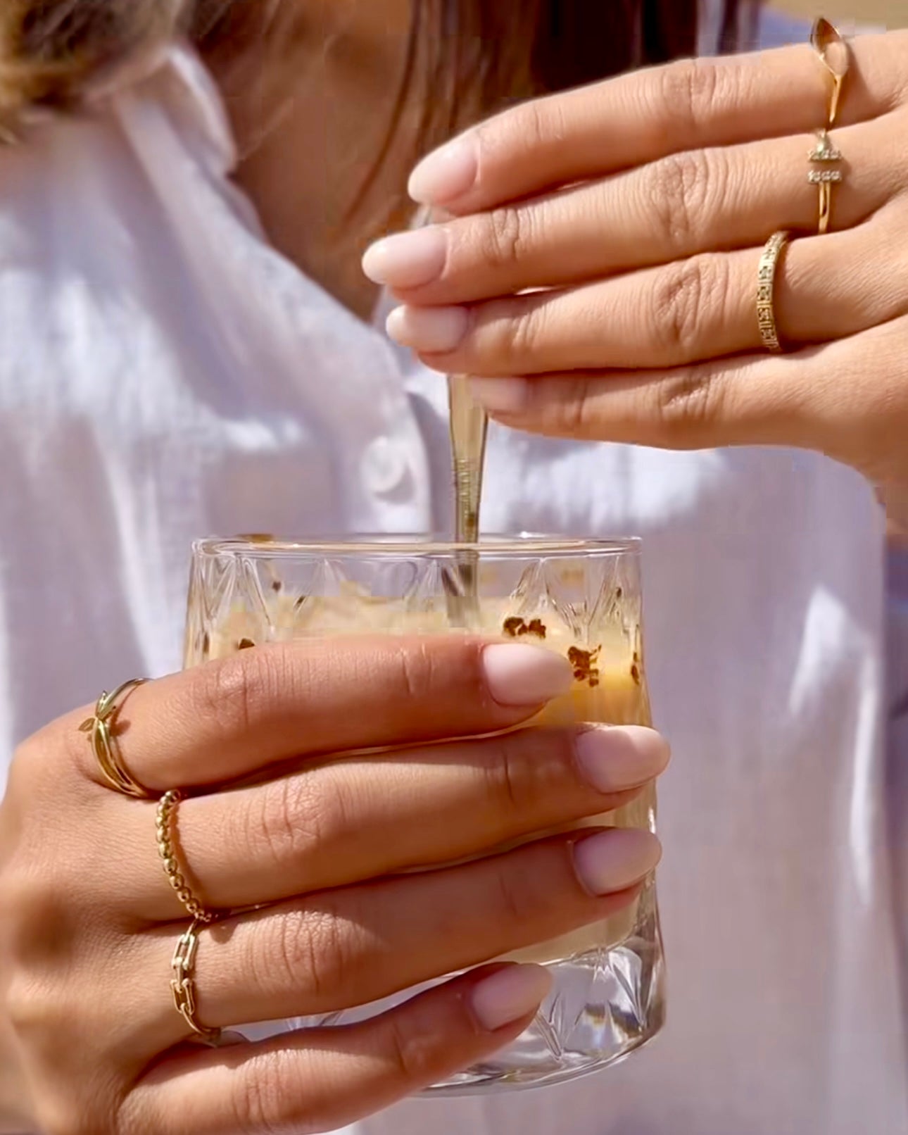 Nahaufnahme zweier Hände mit mehreren filigranen Ringen aus 18 Karat Gelbgold, die ein Glas Eiskaffee mit Löffel halten – verschiedene Ringdesigns von zarten Gliederketten bis zu minimalistischen Symbolen, elegant kombiniert für einen modernen Alltagslook.