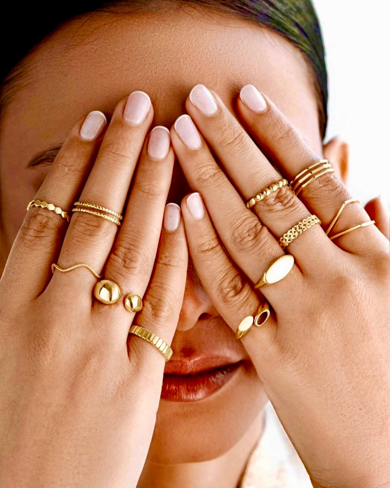 Nahaufnahme einer Frau, die ihr Gesicht mit den Händen bedeckt – beide Hände geschmückt mit zahlreichen Ringen aus 18 Karat Gelbgold in unterschiedlichen Designs: von filigranen, geometrischen und glatten Formen bis hin zu strukturierten und kugelförmigen Elementen. Luxuriöser, moderner Schmuck-Look.