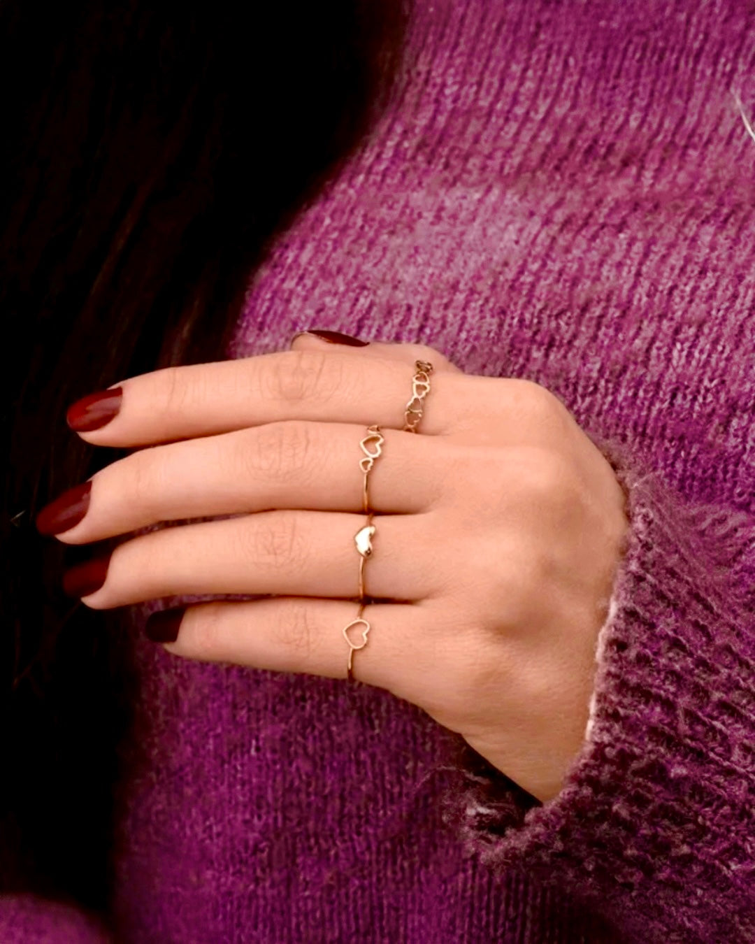 Hand mit bordeauxroten Nägeln trägt mehrere filigrane 18k-Gelbgold-Ringe mit kleinen offenen Herzmotiven, vor einem violetten Strickpullover.