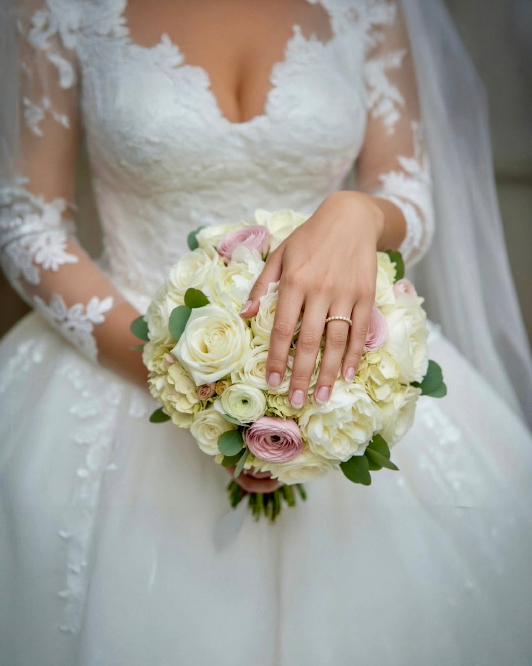 Braut in elegantem Spitzenhochzeitskleid hält einen Blumenstrauß aus weißen und zartrosa Rosen, Nahaufnahme der Hand mit feinem Diamantring, romantische Hochzeitsästhetik.