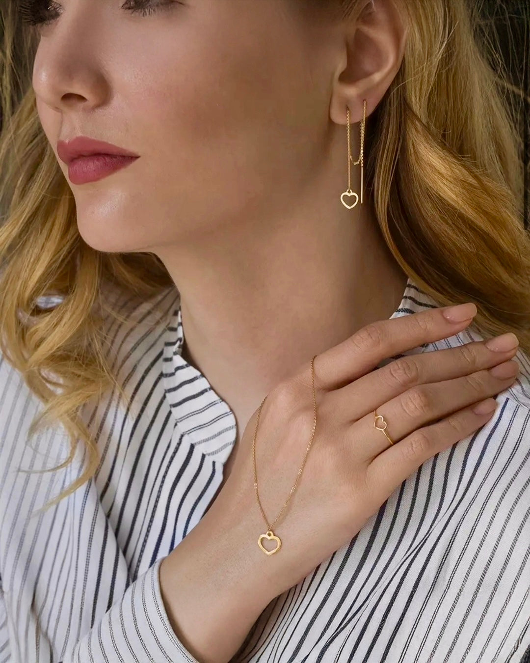 Blonde Frau in gestreifter Bluse trägt ein elegantes Schmuck-Set aus 18-karätigem Gelbgold: zarte Herz-Ohrringe, eine feine Halskette mit offenem Herzanhänger und ein passender filigraner Herzring an der rechten Hand.