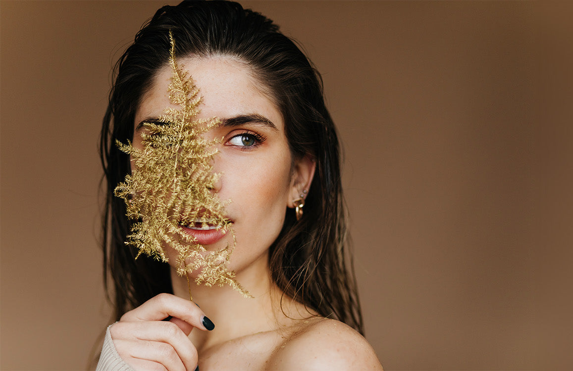 Hübsche junge Frau mit dunklen Haaren, goldener Ohrring, hält eine filigrane, goldüberzogene Pflanze vor die rechte Gesichtshälfte und schaut nach links ins Off.