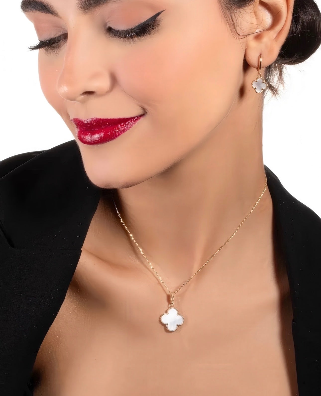 Elegante Frau mit hochgestecktem Haar trägt eine filigrane 18k-Gelbgoldkette mit kleeblattförmigem Anhänger aus weißem Perlmutt sowie passende Creolen mit Perlmutt-Element, vor hellem Hintergrund.