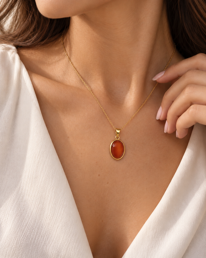Ovaler Anhänger aus 18K Gelbgold mit rot-orangefarbenem Schmuckstein in goldener Fassung, getragen an einer feinen Goldkette am Dekolleté eines weiblichen Models.