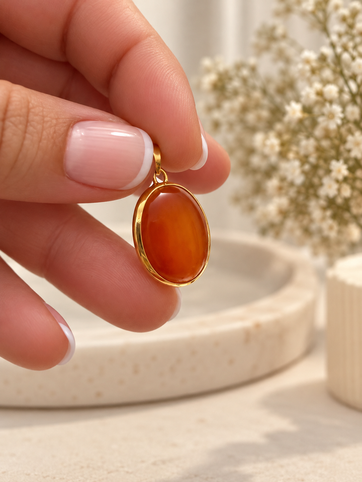 Akhgar Anhänger aus 18 Karat Gelbgold mit ovalem rot-orangefarbenem Schmuckstein, zwischen den Fingern gehalten.