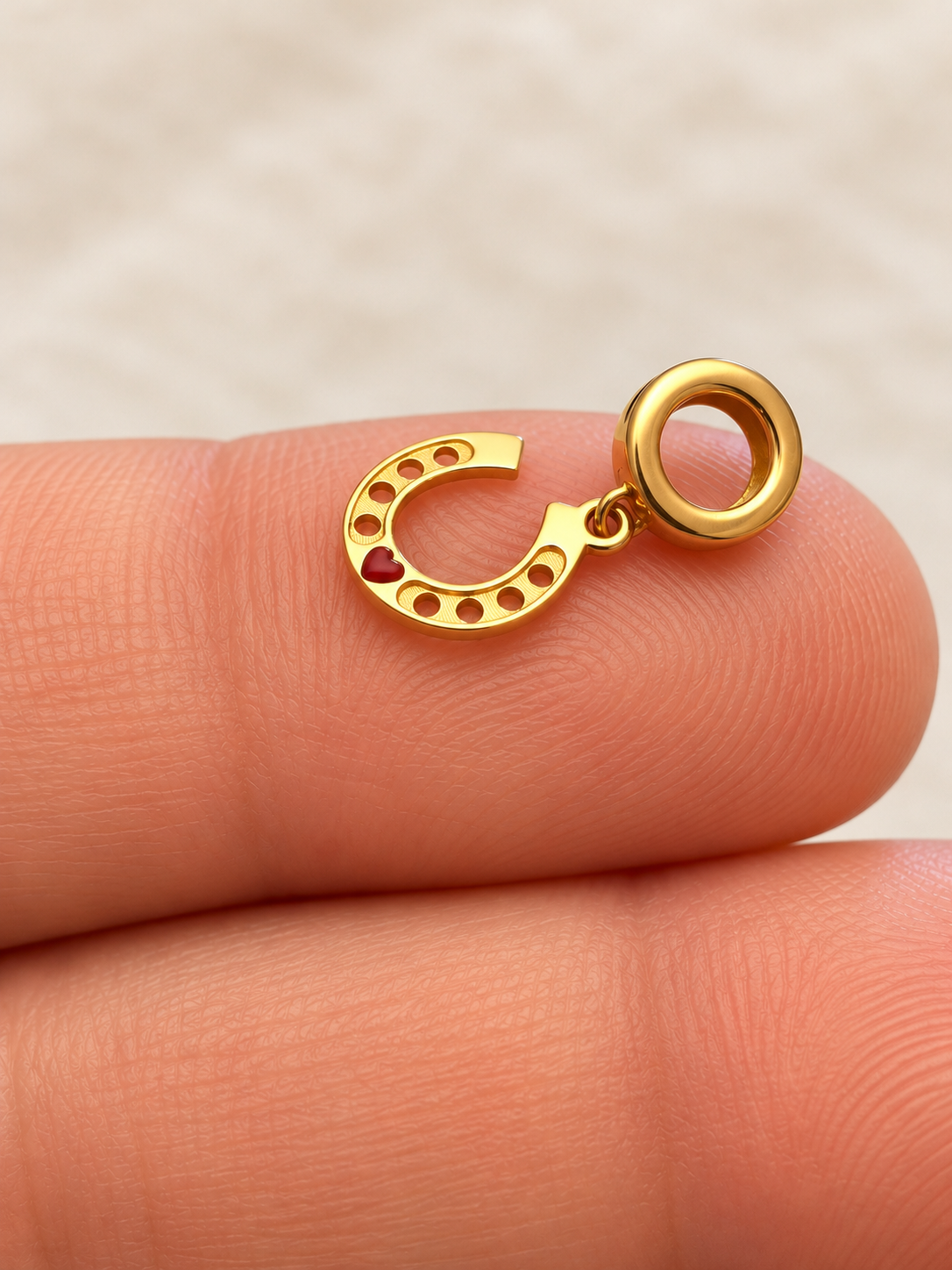 Kleiner Hufeisen-Anhänger aus 18K Gelbgold mit rot bemaltem Herz und runder Öse, seitlich auf einer Fingerspitze liegend.