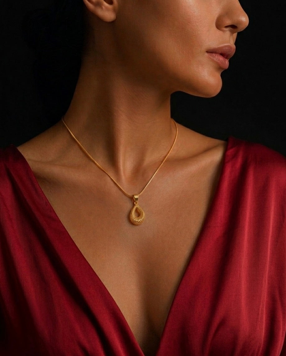 Elegante Frau trägt einen filigranen Anhänger aus 18 Karat Gelbgold im tropfenförmigen Flechtdesign an einer feinen Goldkette. Das kunstvoll gefertigte Schmuckstück liegt harmonisch auf dem Dekolleté und wird durch ein bordeauxrotes Kleid sowie einen dunklen Hintergrund stilvoll in Szene gesetzt. Luxuriöser Damenschmuck mit persischer Handwerkskunst für zeitlose Eleganz.