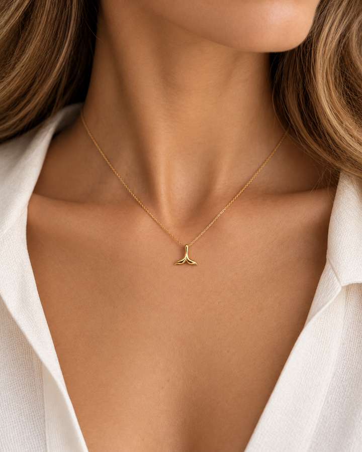 Filigraner Anhänger in Form einer stilisierten Walflosse aus glänzendem Gelbgold, getragen an einer feinen Goldkette am Dekolleté einer Frau mit weisser Bluse.