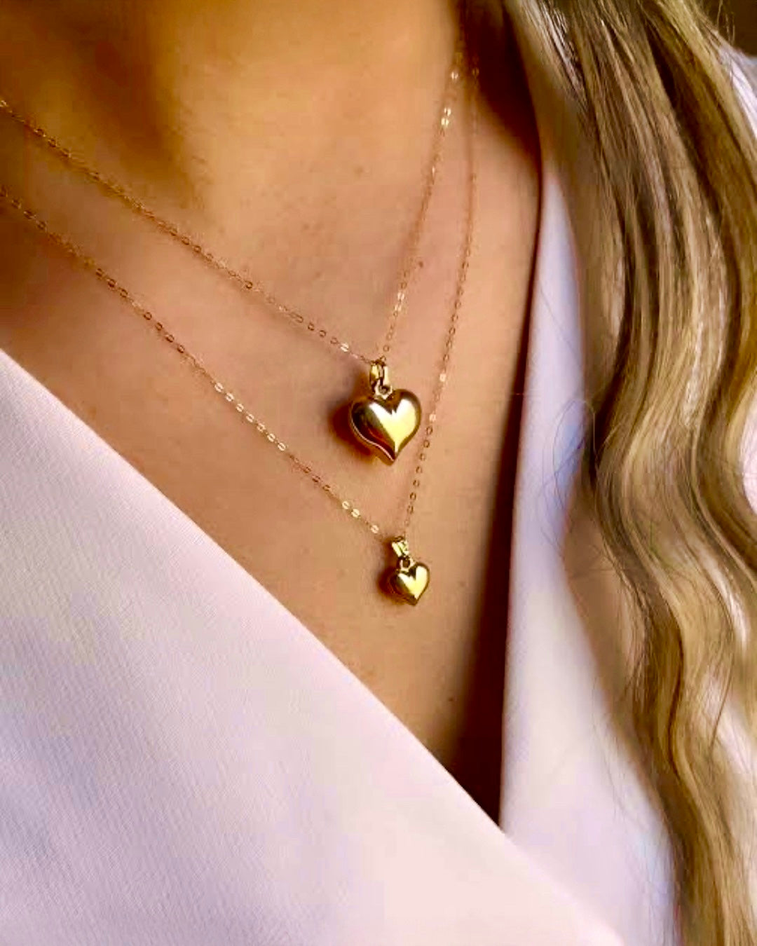 Zwei feine 18k-Gelbgoldketten mit Herzanhängern in unterschiedlichen Grössen, elegant übereinander getragen auf dem Dekolleté; glänzende, hochpolierte Oberfläche, stilvoll kombiniert mit hellem Outfit.
