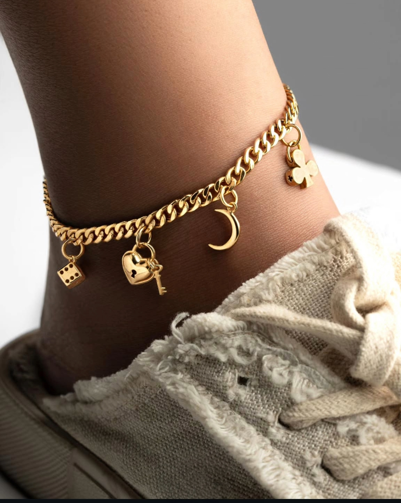Fußkette aus 18 Karat Gelbgold mit verschiedenen Anhängern – darunter ein Würfel, ein Herz mit Schlüssel, ein Mond und ein Kleeblatt – elegant am Knöchel getragen, kombiniert mit hellen Stoffschuhen für einen modernen, verspielten Look.