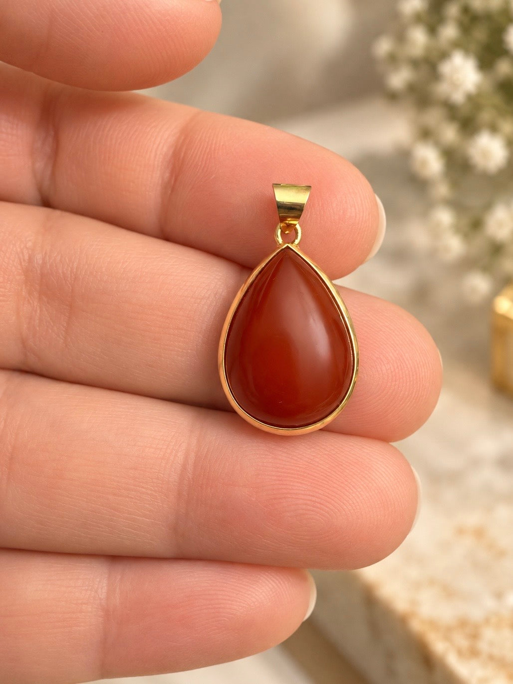 Anhänger aus 18k Gelbgold mit tropfenförmigem, rostrotem Schmuckstein in Nahaufnahme auf den Fingern einer Hand.
