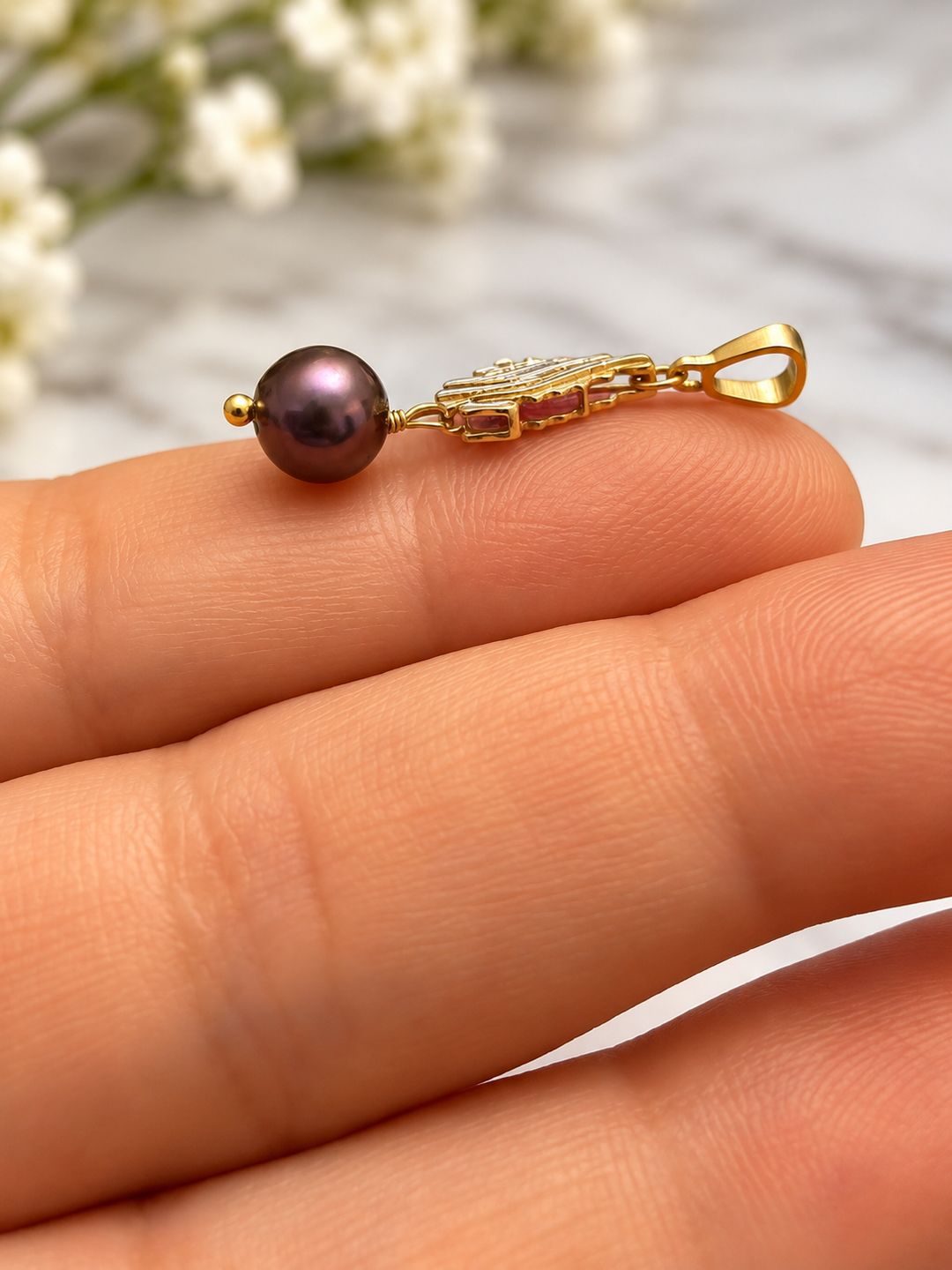 Makroaufnahme eines filigranen Anhängers aus 18k Gelbgold mit dunkelvioletter Zuchtperle und rosafarbenen Details, seitlich auf den Fingern einer Hand präsentiert.