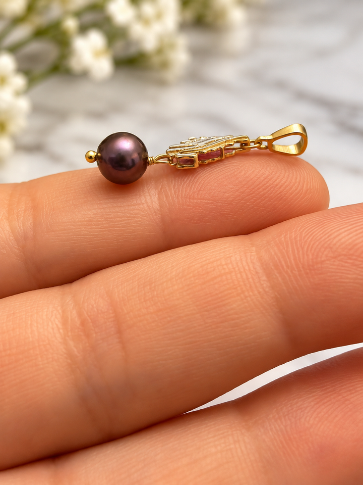 Makroaufnahme eines filigranen Anhängers aus 18k Gelbgold mit dunkelvioletter Zuchtperle und rosafarbenen Details, seitlich auf den Fingern einer Hand präsentiert.