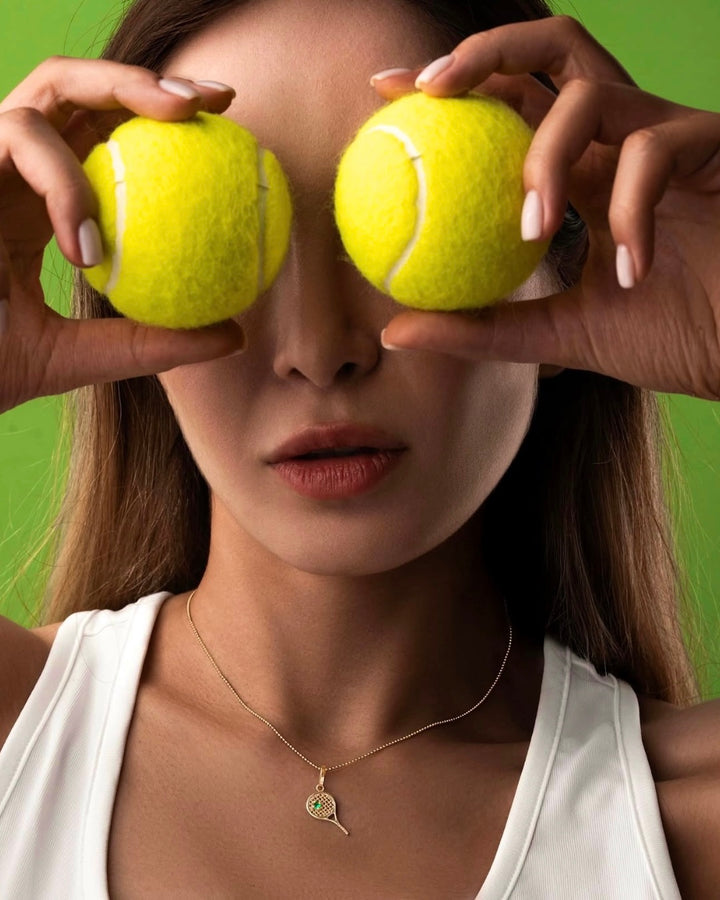 Anhänger aus 18k Gelbgold in Tennisraketen-Form mit grünem Zirkonia, getragen am Hals, Lifestyle-Bild mit Tennisbällen