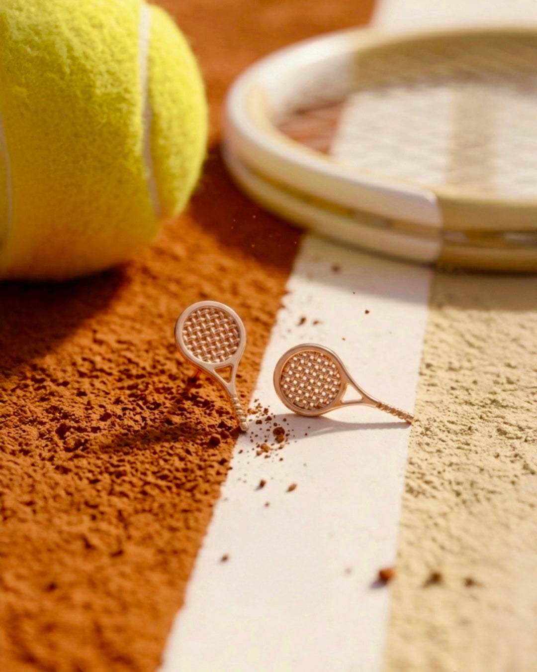 18 Karat Gold-Ohrstecker in Form von Tennis-Schlägern auf einem Tennisplatz aus Sand und Kreide. Die filigranen Schläger zeigen ein detailreiches Besaitungsmuster und werden vor Tennisbällen und einem Tennisschläger kunstvoll in Szene gesetzt. Eleganter Goldschmuck für Tennisliebhaber.