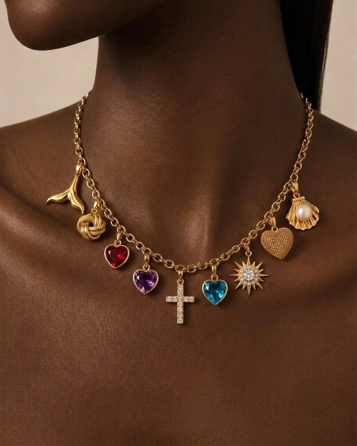 18 Karat Gold Charm-Halskette mit verschiedenen Anhängern an einem Model getragen. Die Kette kombiniert ein funkelndes Kreuz mit Zirkonia, Herzanhänger mit roten, violetten und blauen Edelstein-Optiken, eine Sonne mit Zirkonia, ein strukturiertes Goldherz, eine Muschel mit Perle, einen Knoten sowie einen Ast-Anhänger. Hochwertiger, eleganter Statement-Schmuck für Damen und Herren mit modernem, luxuriösem Design.