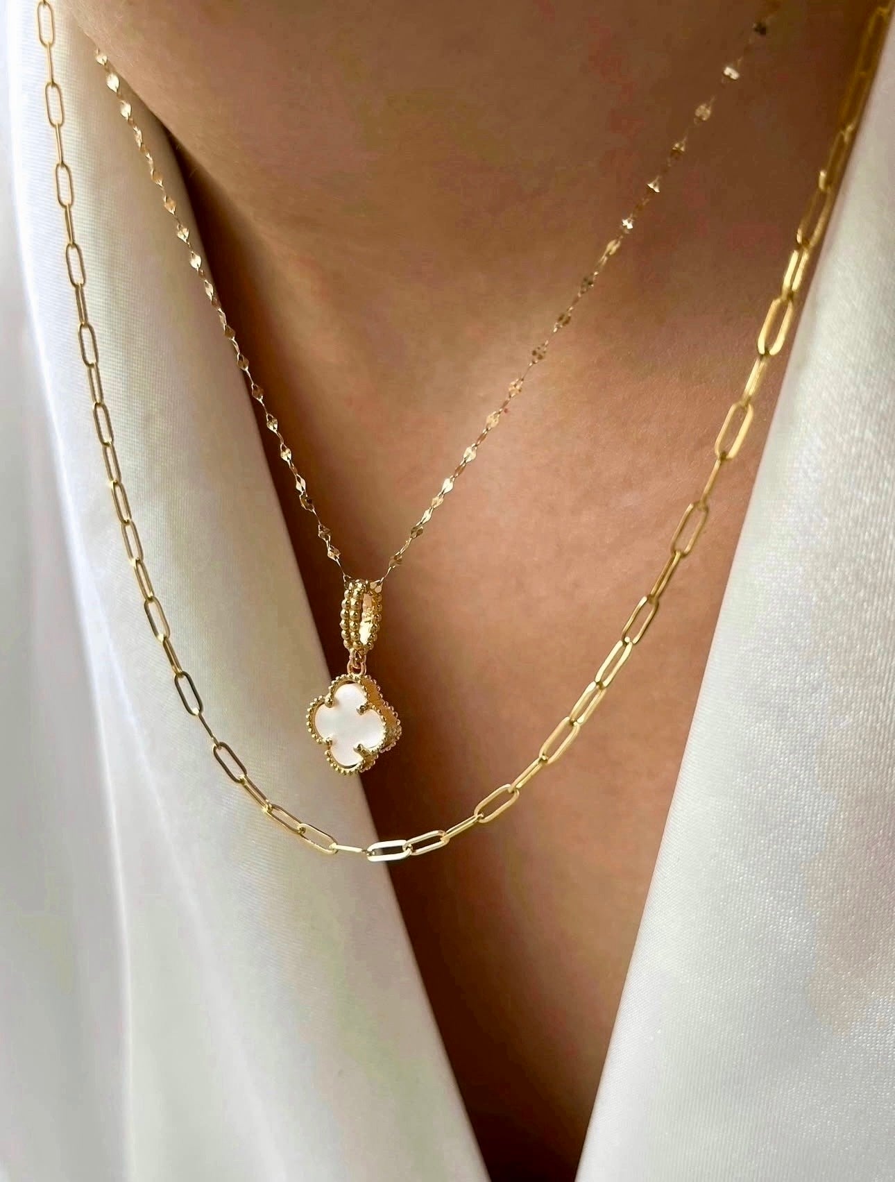 Feine Halskette aus 18k Gelbgold mit einem kleinen, vierblättrigen Glücksanhänger aus weißem Perlmutt, eingefasst in eine verzierte goldene Rahmung. Der Anhänger hängt an einer filigranen Goldkette und wird kombiniert mit einer zweiten, etwas gröberen Gliederkette aus 18k Gelbgold.