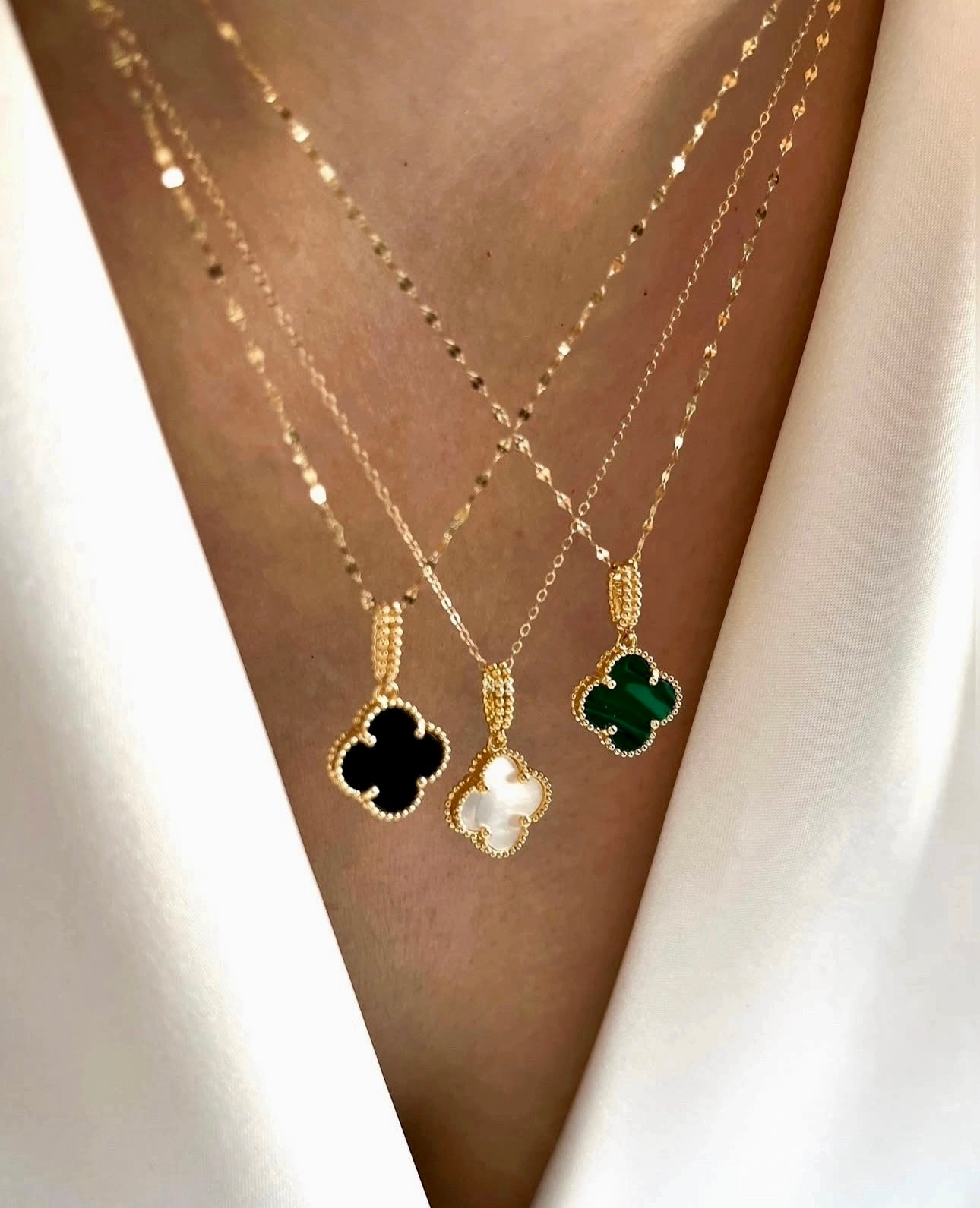 Anhänger in vierblättriger Form aus weißem Stein, eingefasst in feine goldene Perlenfassung, an einer zarten Goldkette, elegant auf heller Stoffbluse getragen.