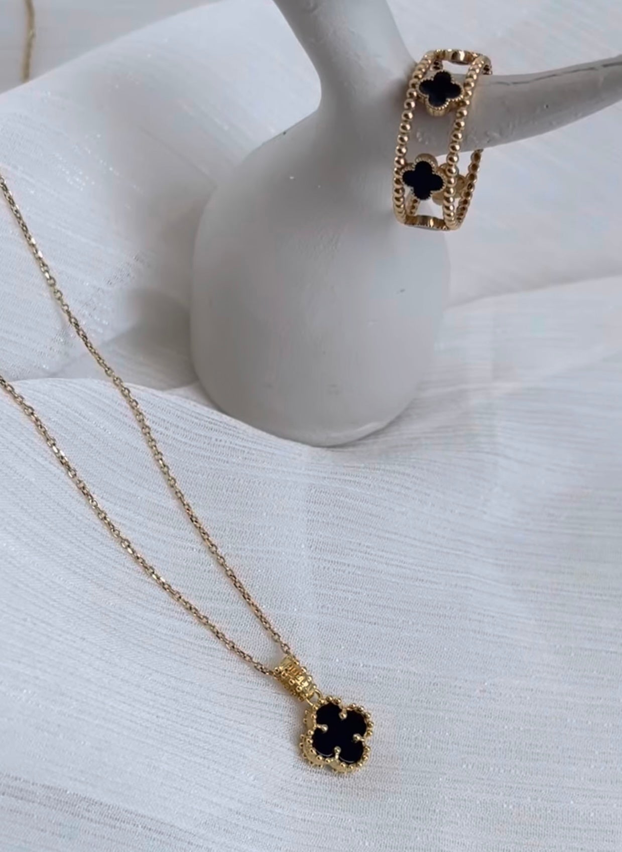 Eleganter 18k Gelbgold-Anhänger in Kleeblattform mit schwarzem Inlay und perlenartigem Goldrand, an feiner Goldkette, inszeniert neben passendem Ring in Schwarz-Gold auf weißem Stoffhintergrund.