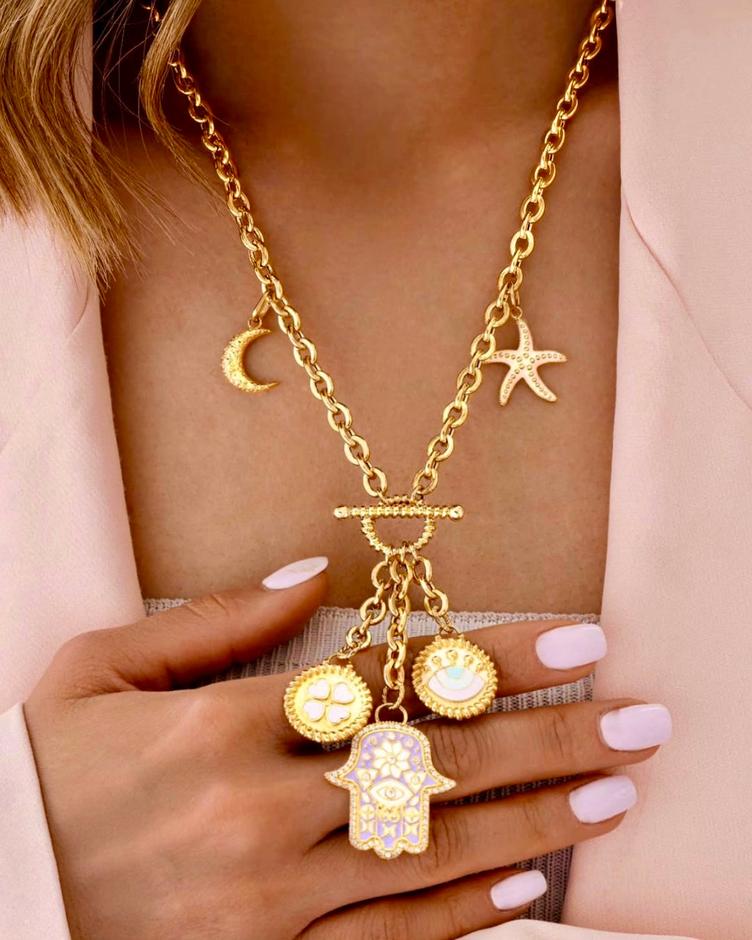 Halskette aus 18 Karat Gelbgold mit auffälligen Anhängern – darunter ein Halbmond, ein Seestern, ein vierblättriges Kleeblatt, ein Symbolauge und eine verzierte Hamsa-Hand – getragen zu einem roséfarbenen Blazer, eleganter und moderner Look mit spiritueller Note.