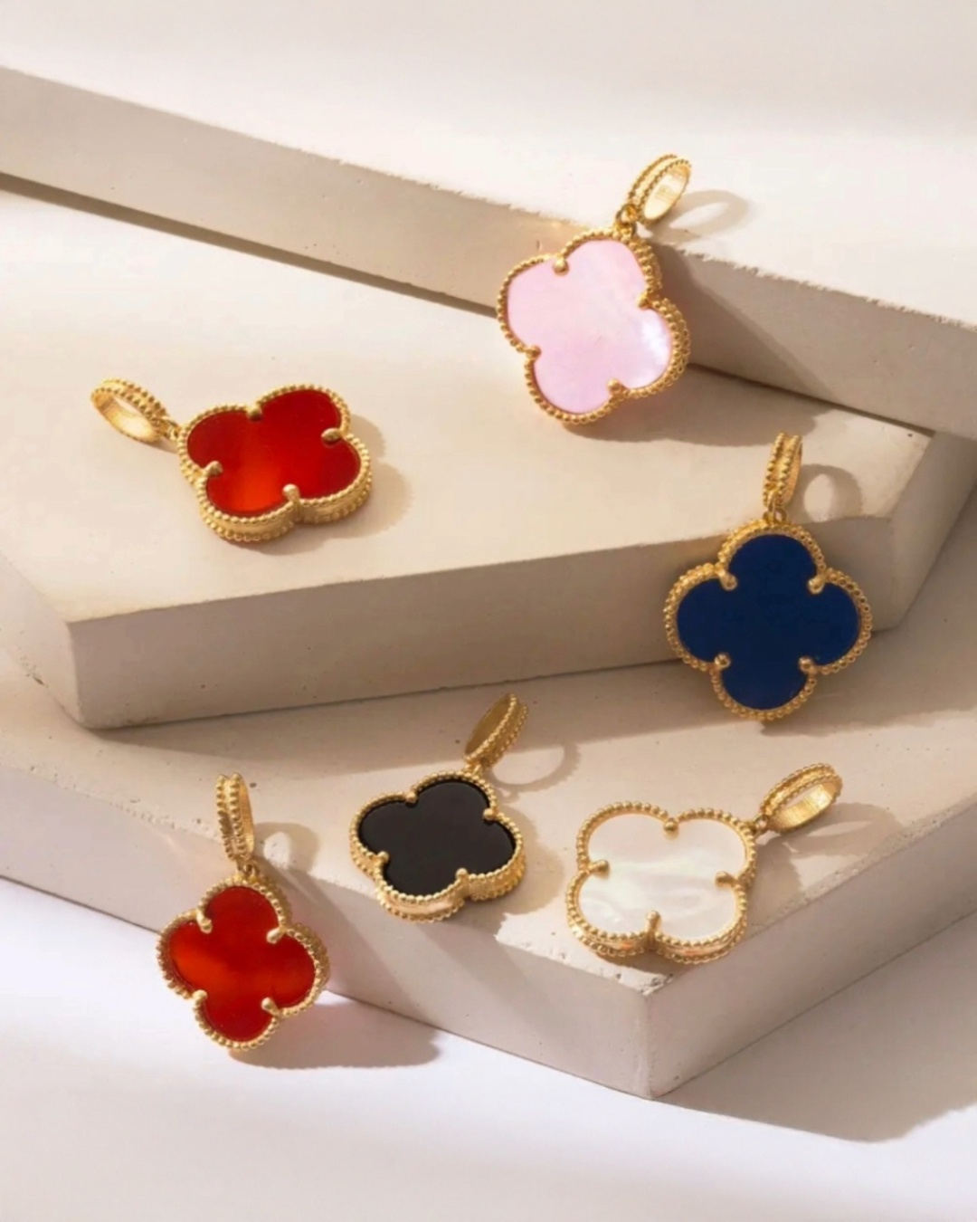 Mehrere Kleeblatt-Anhänger aus 18k Gelbgold mit fein gearbeiteter Perlenfassung, präsentiert in verschiedenen Farbvarianten – Rosa, Rot, Blau, Schwarz und schimmerndes Weiss – elegant arrangiert auf hellen Podesten, luxuriöser Schmuck mit edlem Stein- oder Perlmutt-Inlay.